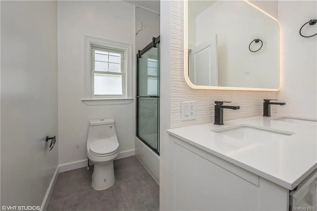 A clean, minimalist design that maximizes both function and style. This hallway bathroom renovation includes:
A floating double-sink vanity with a bright white quartz top
Sleek matte black fixtures and matching hardware for a bold, modern contrast
A stunning backlit LED mirror that adds elegance and soft ambient lighting
Frameless glass tub/shower enclosure for a contemporary, open look
Large-format stone-look floor tile for durability and sophistication
Simple, neutral color palette for a timeless spa-like feel